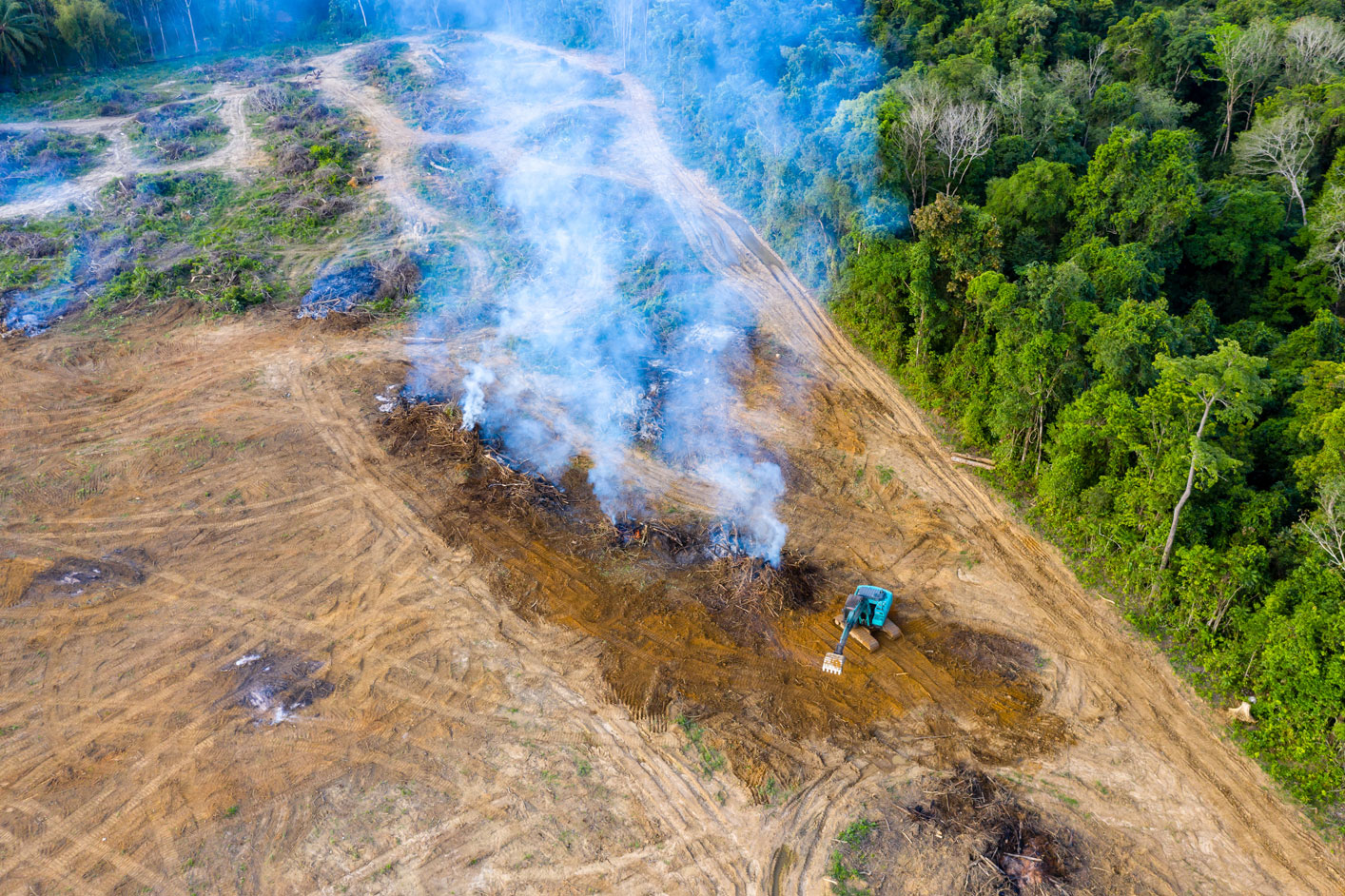 Deforested land with smoke and a small digger with a small area of green forest