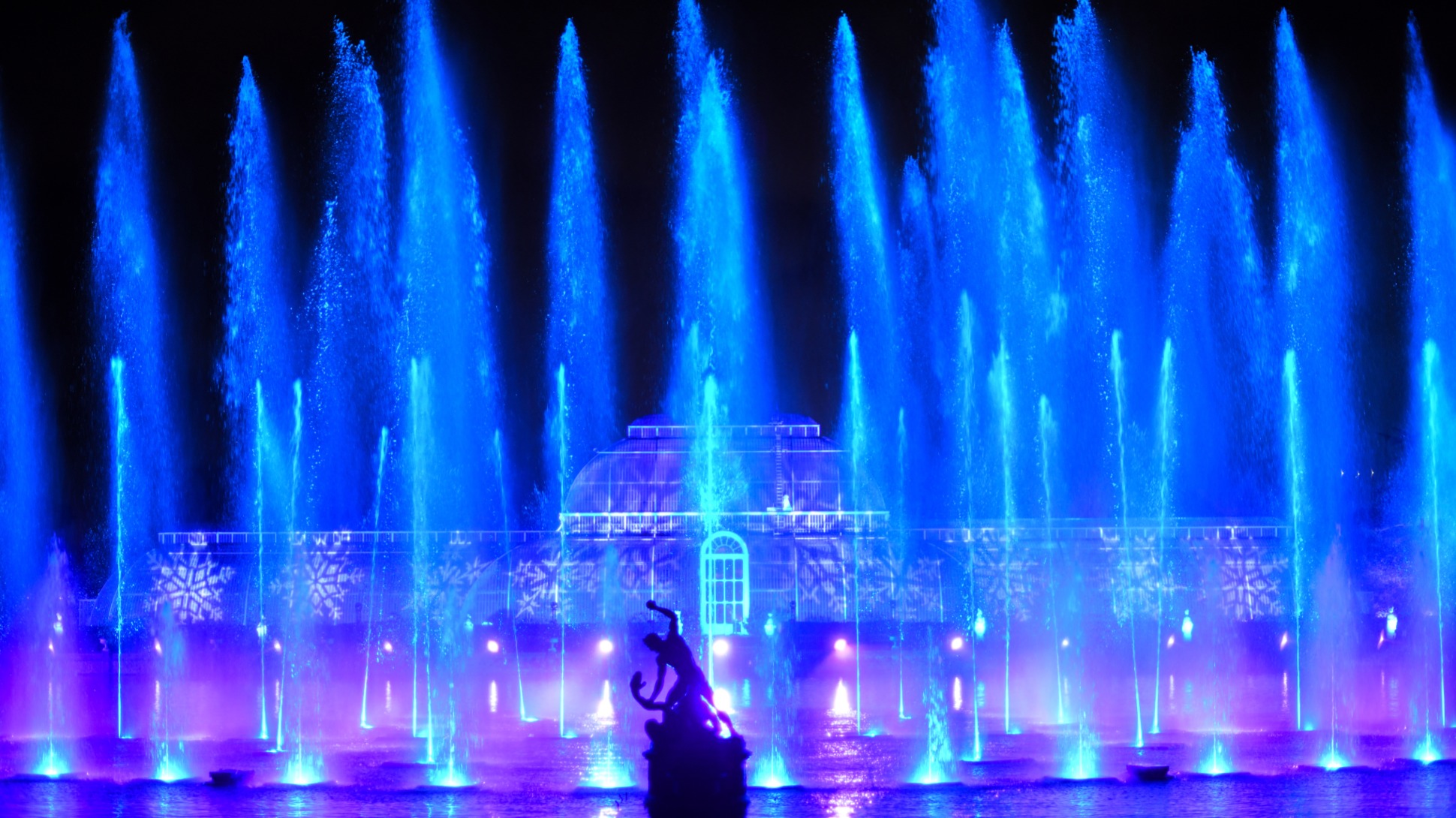 Palm House lit up blue and purple, vertical fountains in foreground