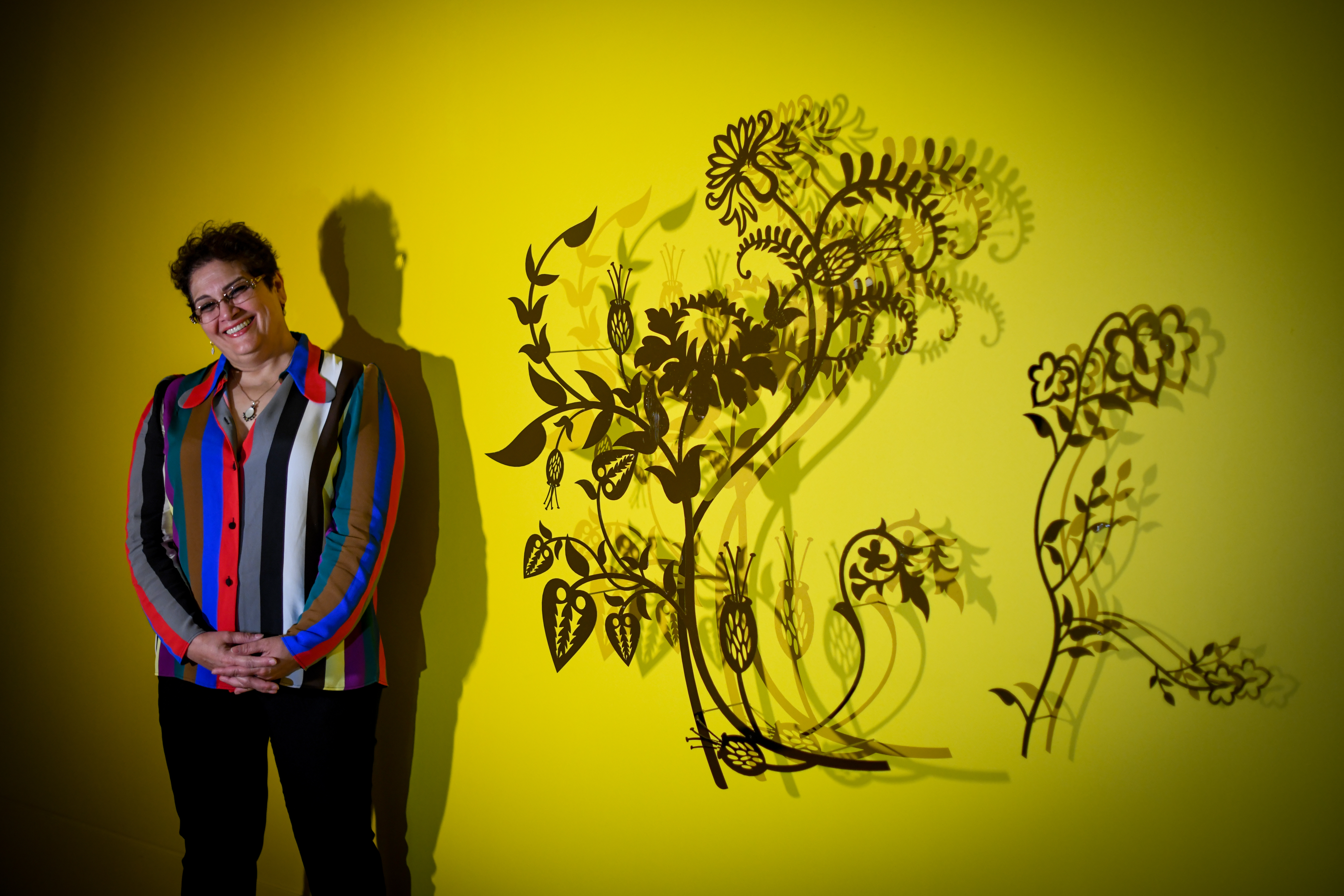 A Pakistani woman with short hair in a striped blouse stands smiling against a yellow wall, next to a dramatically lit wall installation of metal flower silhouettes which cast shadows on the wall
