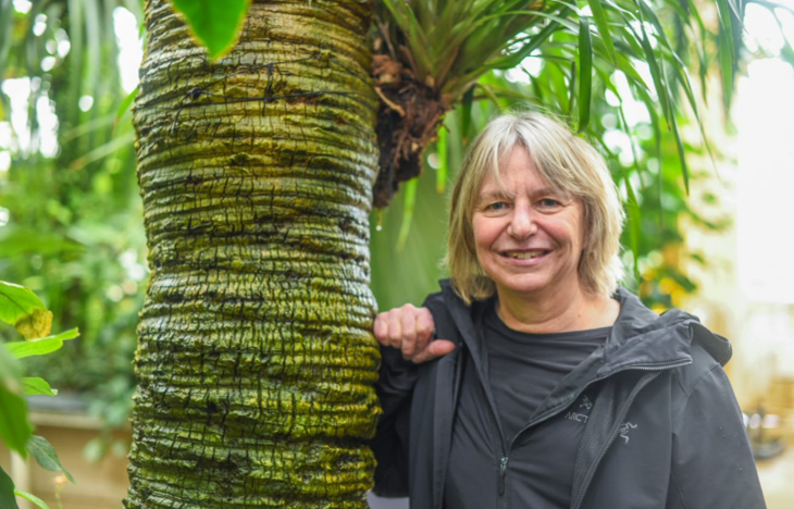 An older white woman with short blonde hair leans on a tree trunk and smiles at the camera