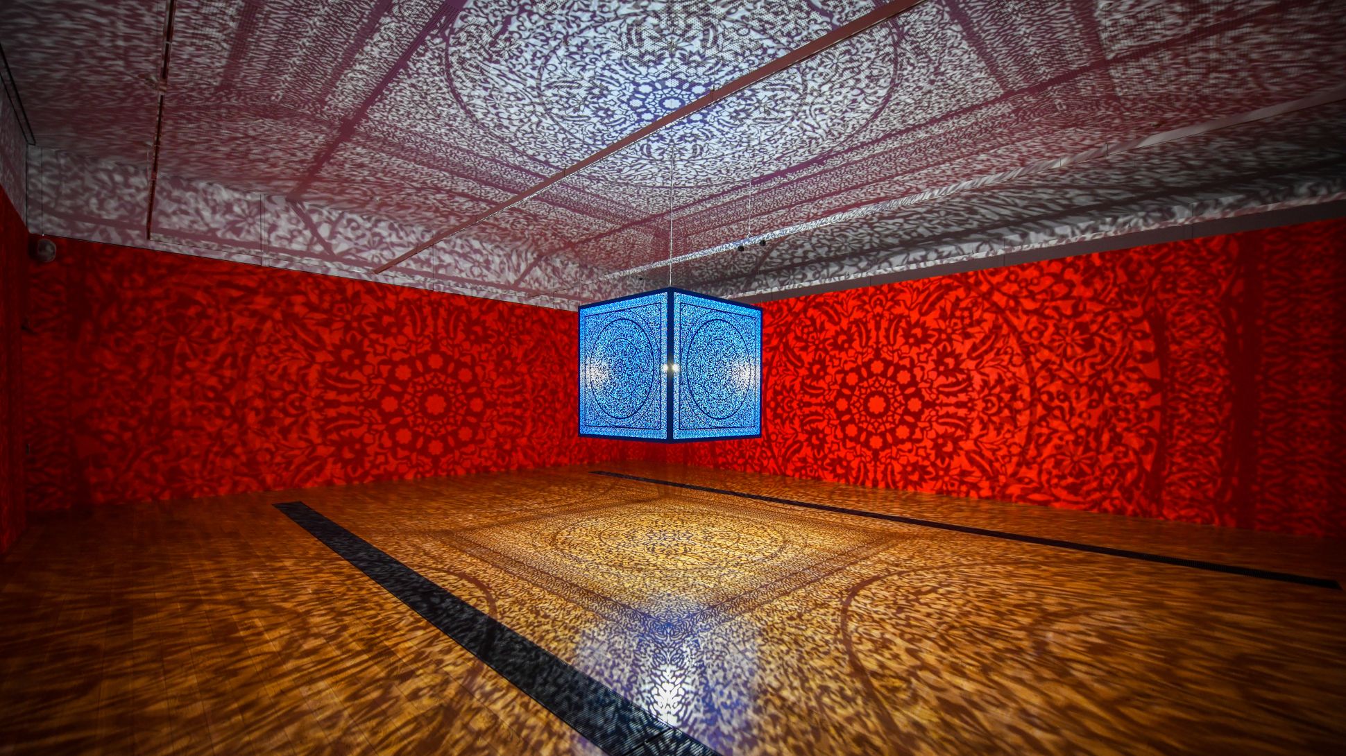 Wide shot: a blue glowing cube hangs suspended in a gallery, throwing intricate shadows onto the orange walls and wooden floor