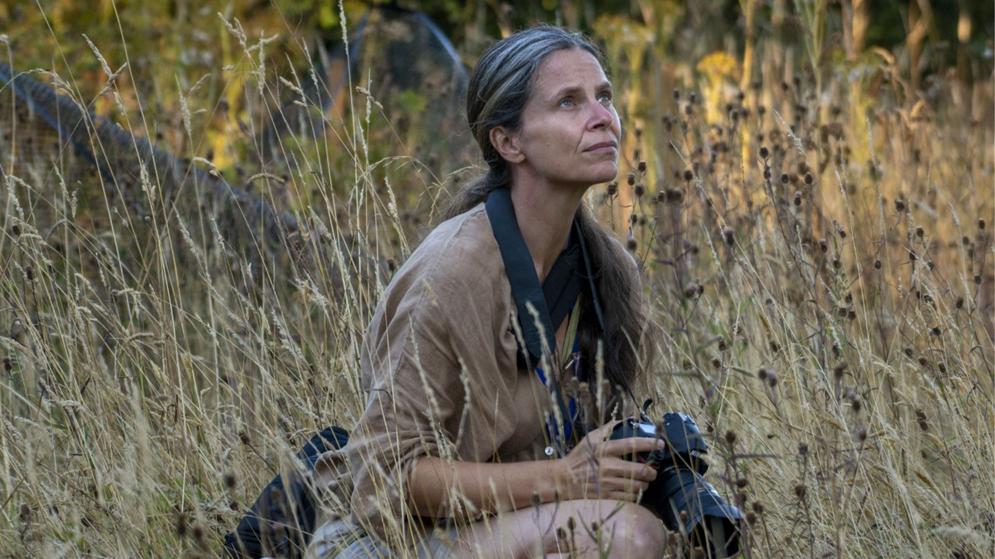 A women is sitting on the ground surrounded by grasses, she is holding a camera and looking up