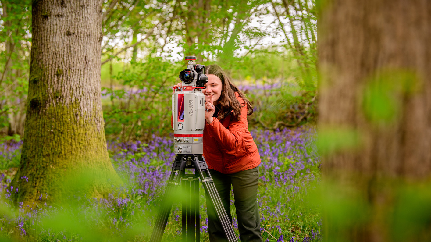 A scientist stands in a field of bluebells, next to a LiDAR scanner