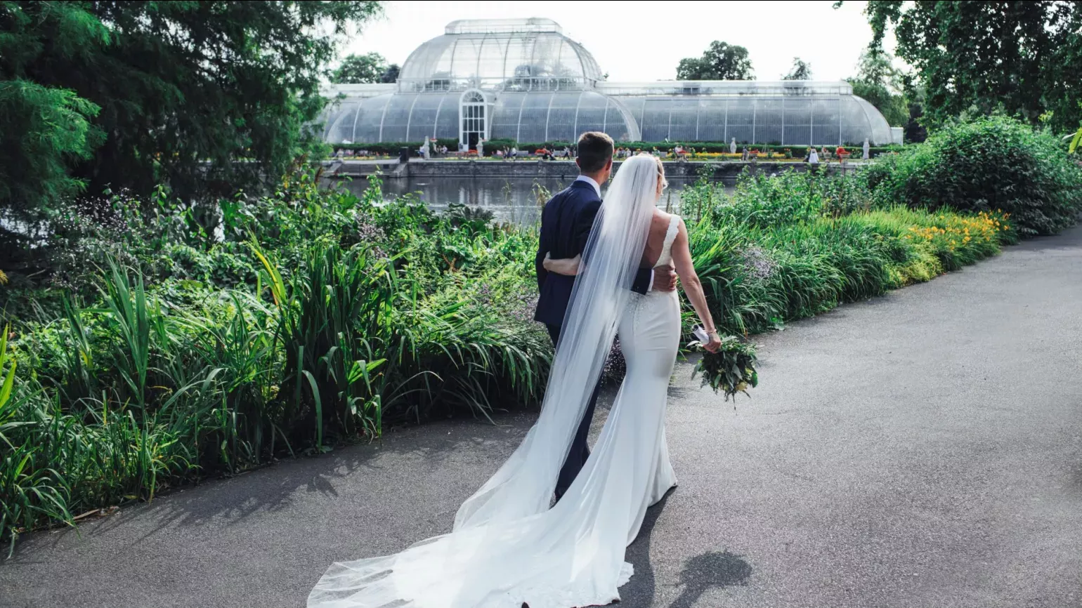 A wedding couple walk along a path in front of the Palm House at Kew Gardens