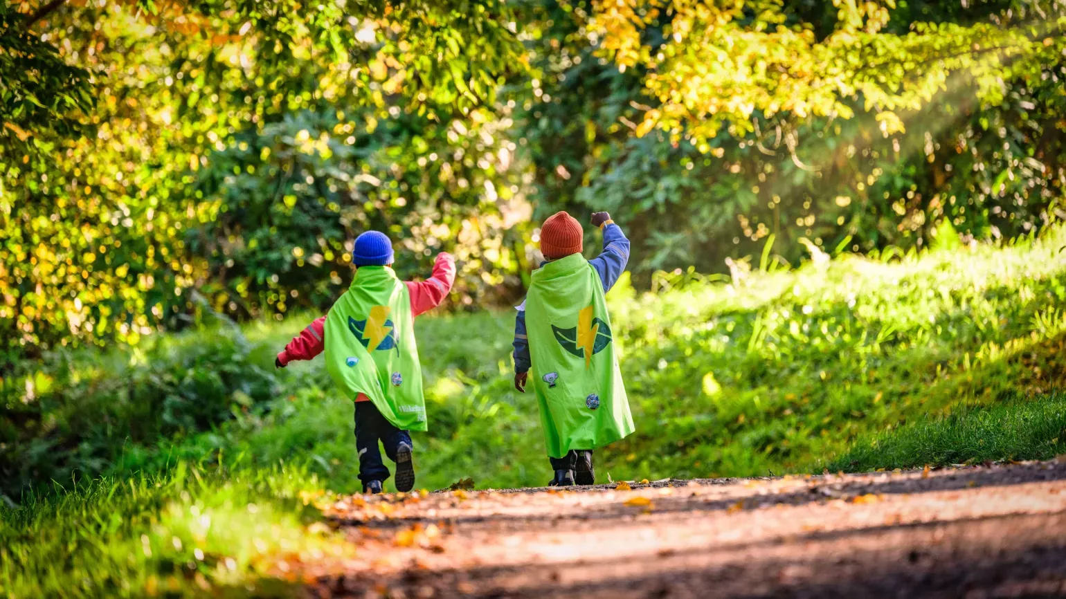 Seen from behind, two children run down a sunny woodland path in autumn, wearing red and blue beanies and green Nature Heroes capes