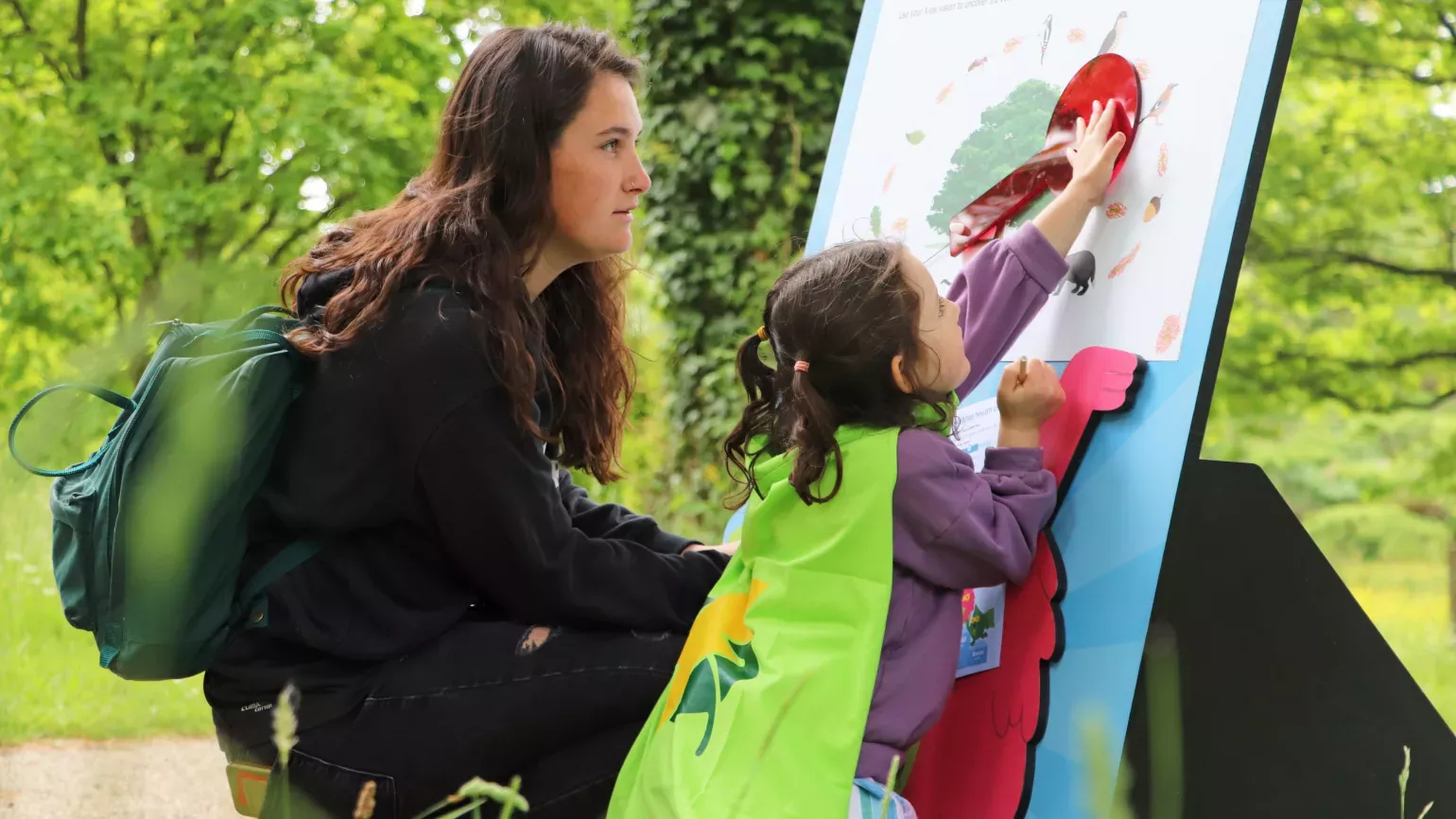 A mother watches her toddler daughter, with pigtails and a green Nature Heroes cape, plays with an interactive life cycle illustration