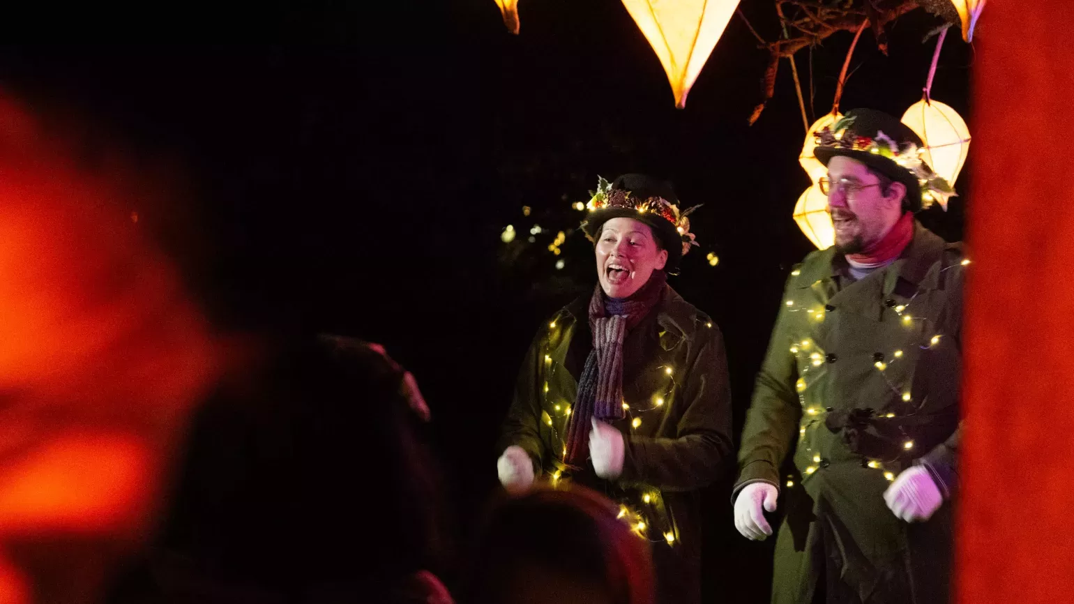 Two choral singers in old fashioned trench coats and bowler hats, festooned with fairy lights