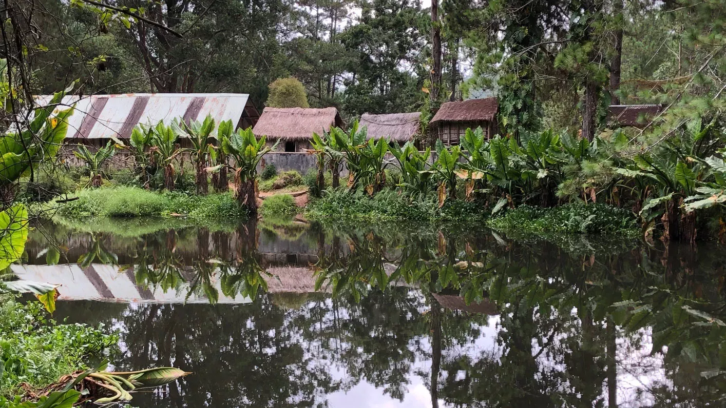 Four huts next to a pond in a green tropical forest in Madagascar. The reflection of the huts and plants can be seen in the pond.