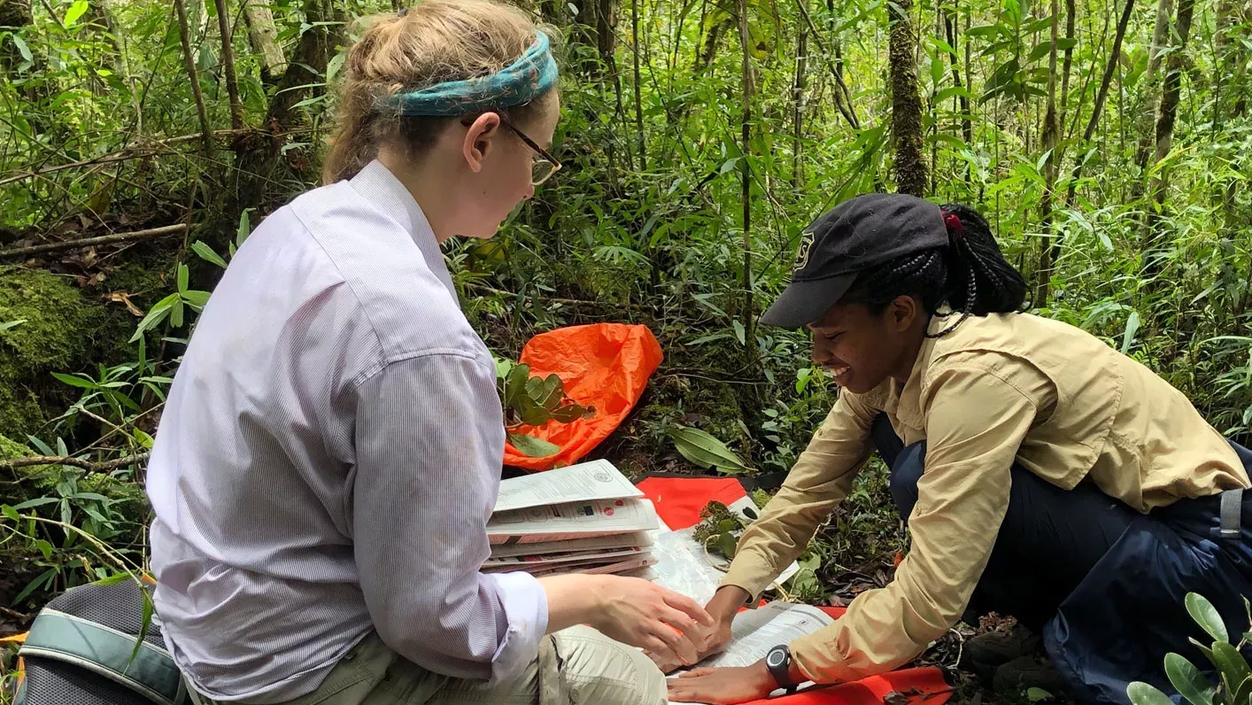 Two students crouched down in a forest. One student is smiling while pressing a plant specimen