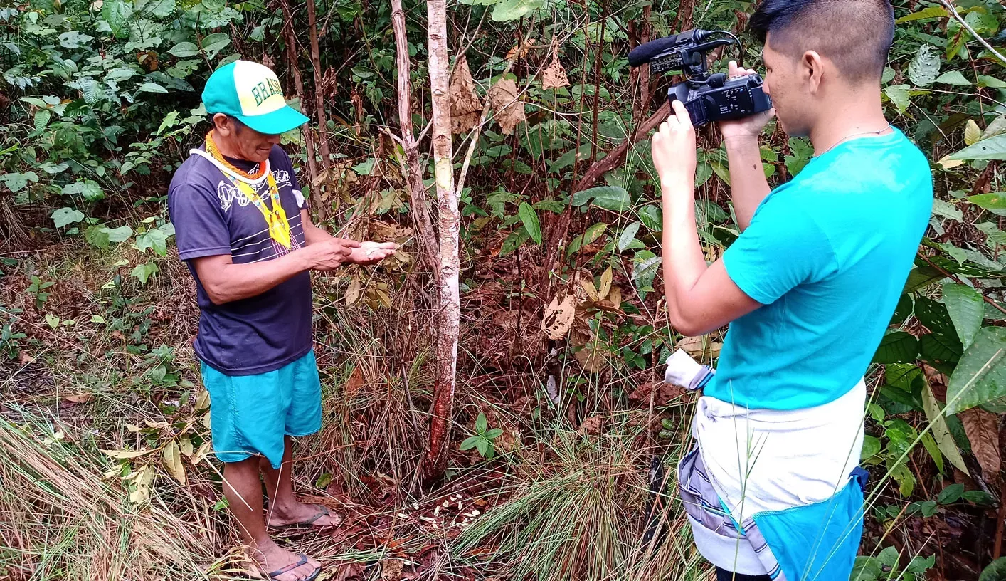Two people in the Amazon rainforest. One person is filming the other with a small film camera who is looking down at the palm of his hand which contains a small plant specimen.