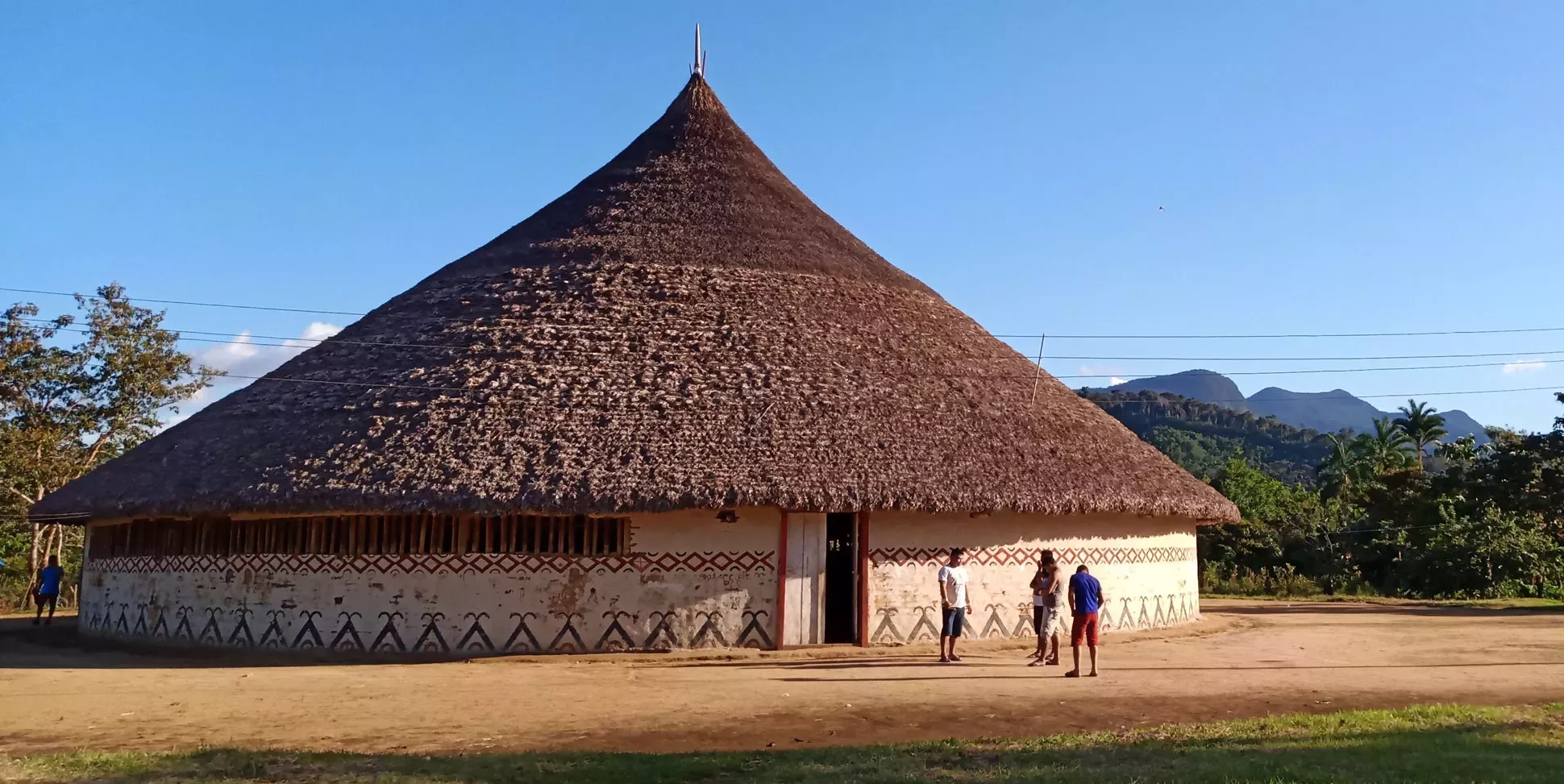 Large round stone hut with a thatched conical roof. Three people are stood in front and look very small in comparison. The sky is blue and the ground is dusty