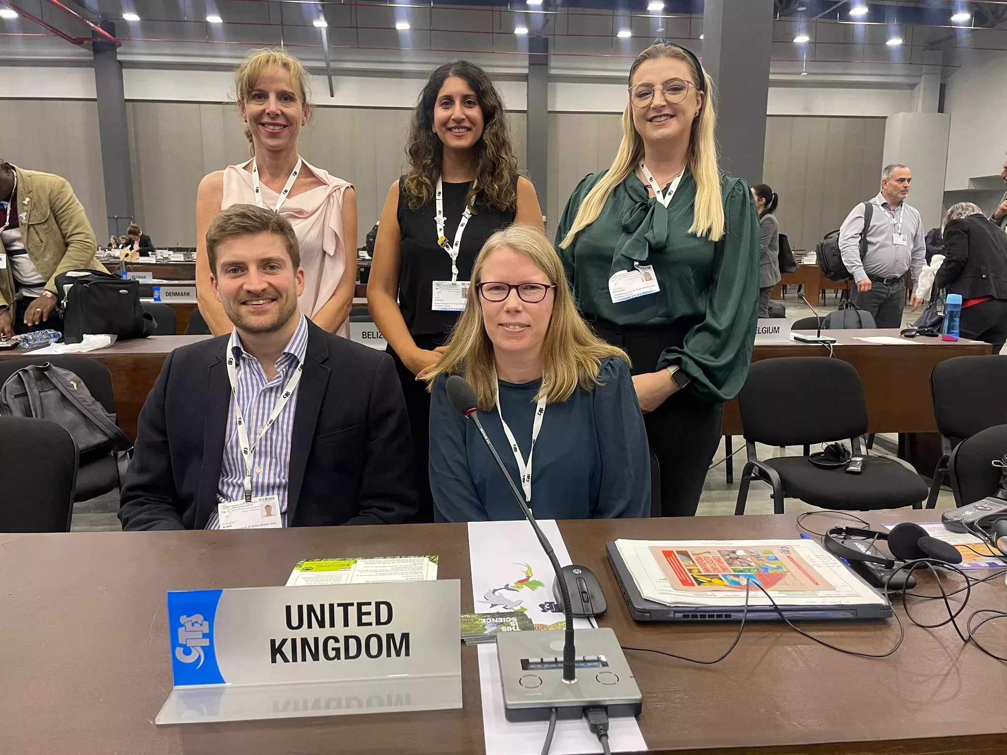 Kew researchers and colleagues pose for a photo at a desk. On the desk are CITES documents, a microphone and a plaque marked United Kingdom