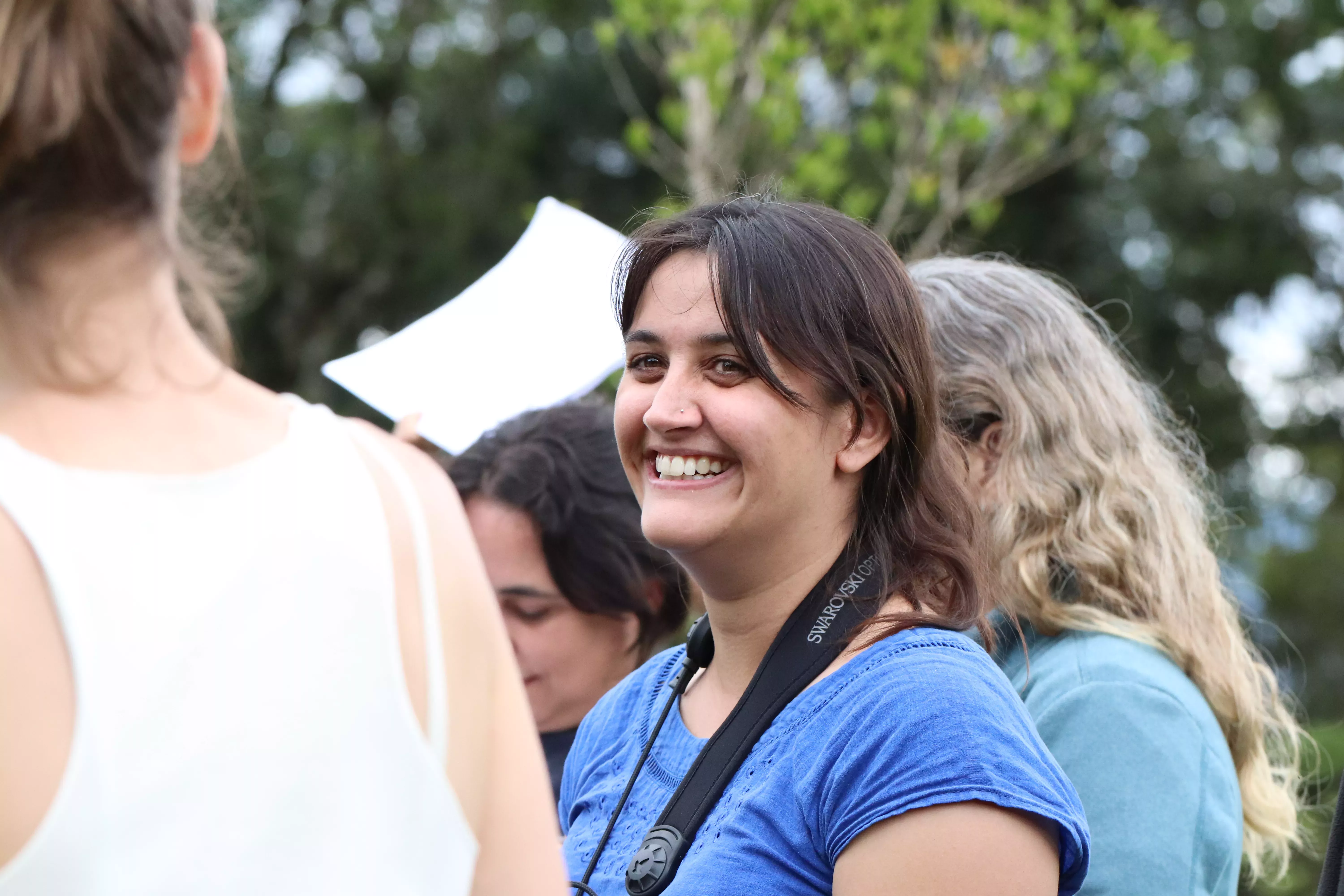 A young Indian woman smiling in a group of other women, looking past the camera, wearing a blue t-shirt and a camera strap. Lush trees in the background.