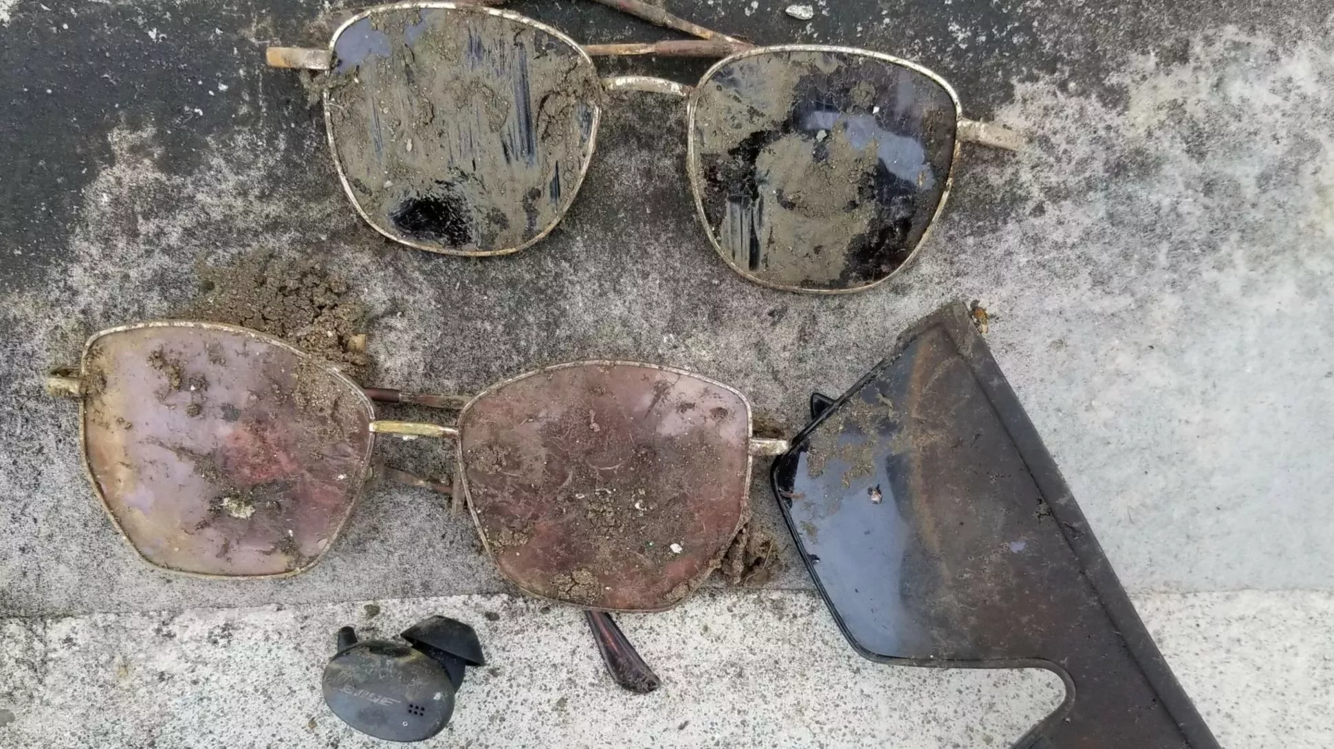 A collection of sunglasses and one Bose earbud covered in mud